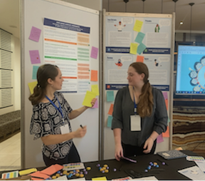Two women are standing in front of a large white board with sticky notes on it speaking