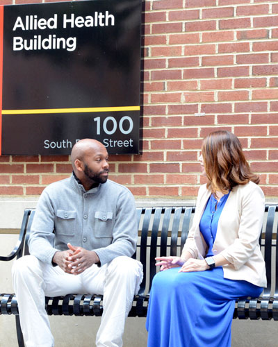 Two people having a conversation on a bench in front of the Allied Health building