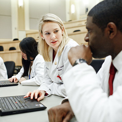 Two students wearing white lab coats in class with laptops