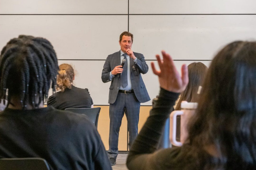 Dean Gladwin speaking in front of a classroom of students with raised hands.