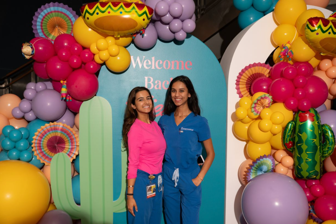 Two medical professionals in scrubs posed in front of a colorful cactus and balloon backdrop.