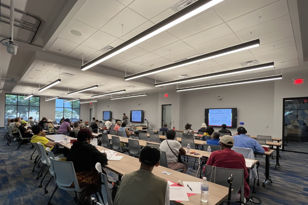 A group of diverse, adult students sitting in a modern classroom facing a lecturer giving a presentation.