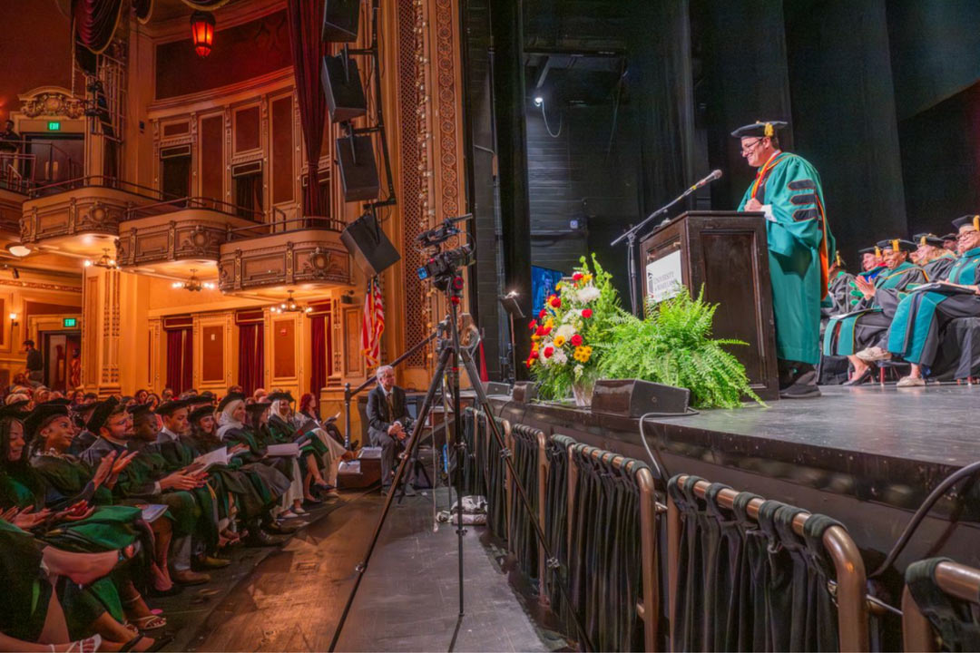 Dean Gladwin speaking in front of theatre in graduation regalia at the Class of 2026 commencement ceremony.