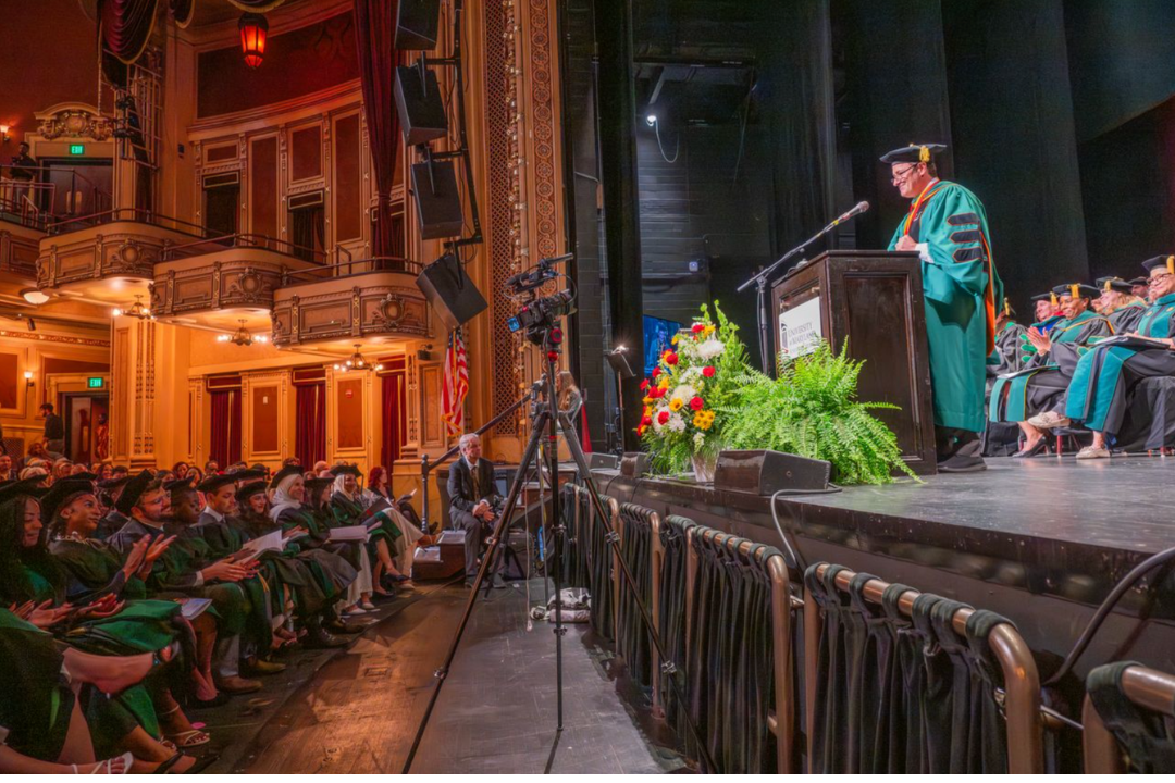 Dean Gladwin speaking in front of theatre in graduation regalia at the Class of 2026 commencement ceremony.
