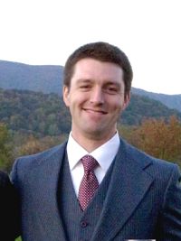 Photo of Collin Brinkman in a navy suit with white shirt and red tie. There are mountains and trees in the background.