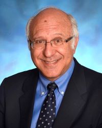 Photo of Dr. Simeon Taylor against a blue background wearing a black suit with blue shirt and blue tie