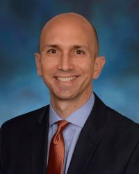 Matthew Laurens, PhD in a black suit with blue shirt and red tie against a blue background