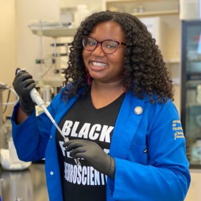 Photo of black woman with curly hair and glasses. She is wearing a black tshirt that says 