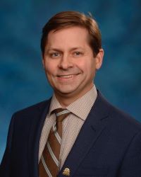 Dr. Todd Gould in a black suit with brown shirt and tie against a blue background