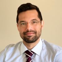 Marcel Lanza, PhD wearing a white shirt and maroon and white striped tie against a beige background