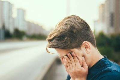 Picture of man with brown hair with his hands pressed against his face. There is a blurry city skyline in the background.