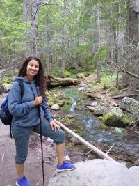 Georgia Rogers in a blue hoodie and pants holding a hiking stick standing in front of a wooded area with a stream