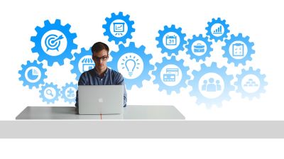 A man sitting at a desk with a laptop open. There are a series of blue gears in the background