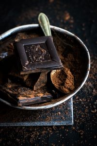 A square of chocolate in a metal bowl full of cocoa powder and a silver spoon. The background is black with more sprinkled cocoa powder.