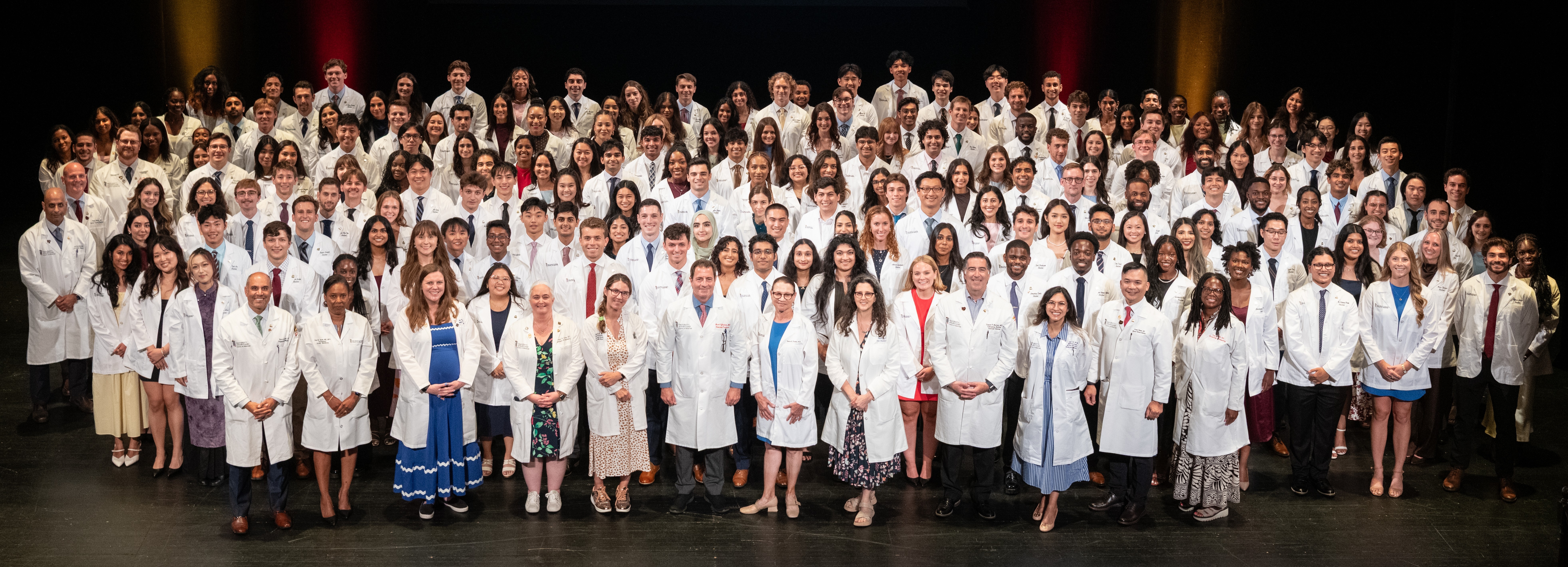 A large group of students and faculty wearing white medical coats posing for a class photo
