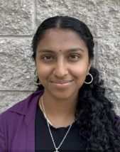 Student headshot. Woman standing in front of a stone wall wearing a black shirt with a purple jacket.