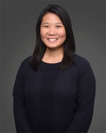 Woman standing in front of a dark grey background wearing a black shirt. Photograph is taken from the waist up.