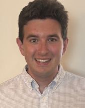 Student headshot. A young man wearing a white button-down shirt, smiling, standing in front of a white wall.