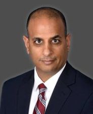 Faculty headshot. Man in a black suit jacket, white shirt, and red tie in front of a grey background.