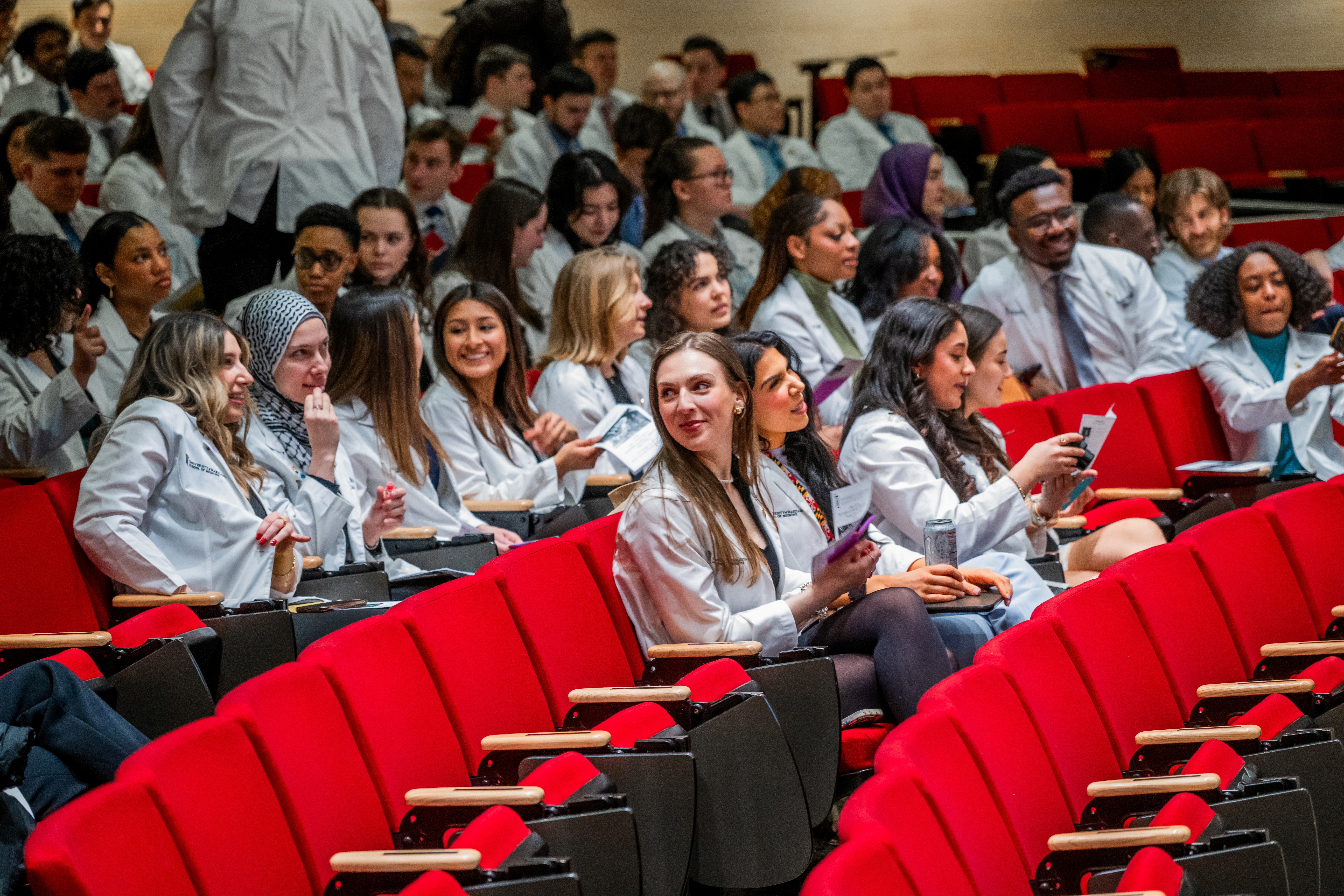Students sitting in red auditorium seats chatting while they await the start of the ceremony